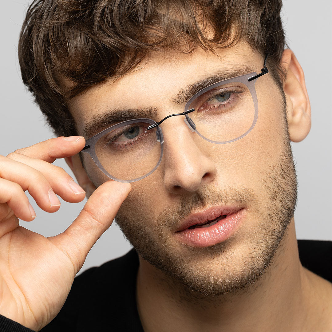Close-up portrait of a man wearing translucent gray, square eyeglasses with a black bridge detail, lightly touching the frame with his hand against a light gray background.